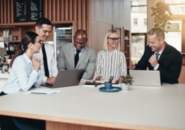 A Group of diverse Supply Chain Manager businesspeople standing together at a table during a meeting in a modern office going over paperwork and working on laptops